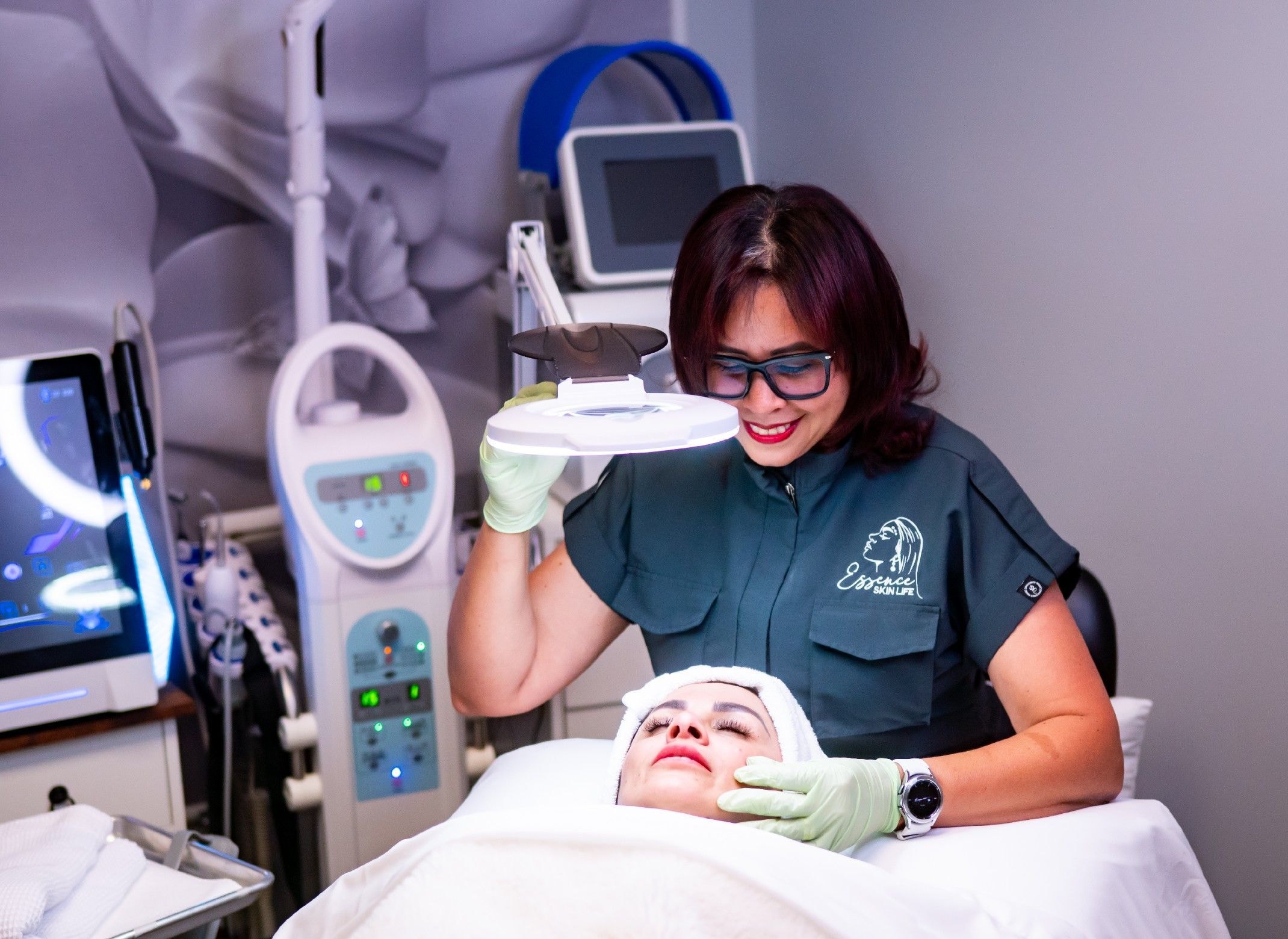 Karina in her Essence uniform examines a client's skin under a magnifying lamp