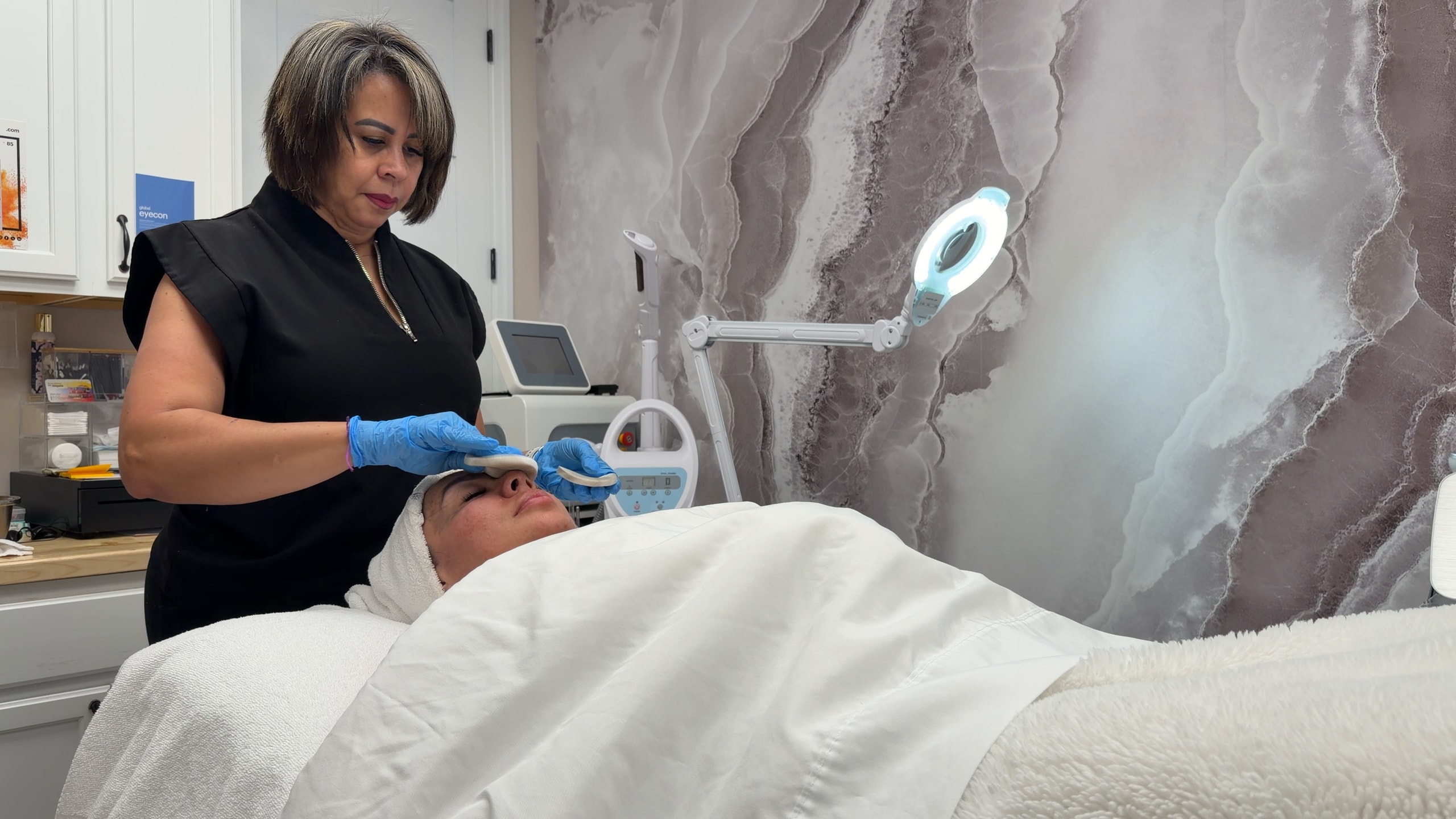Karina cleans a client's face beside the magnifying lamp in the treatment room