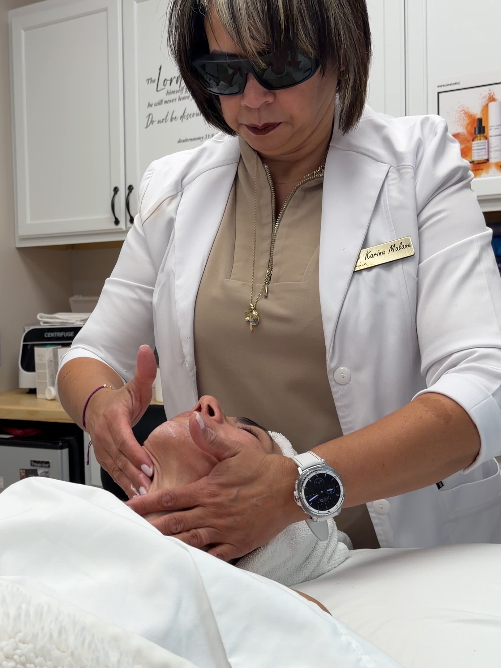 Karina begins a facial, her hands resting on the client's face